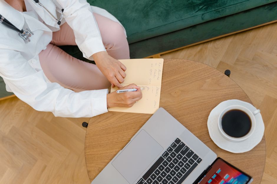 Overhead view of a doctor writing notes during a medical consultation with coffee and laptop on the table.