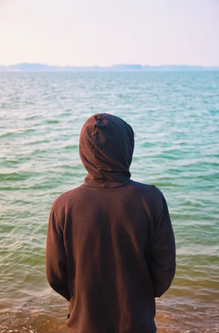 Person wearing a hoodie stands peacefully by a serene lake, enjoying leisure and relaxation.
