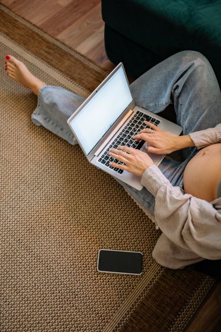 Pregnant woman sitting on carpet using a laptop at home, embracing technology.