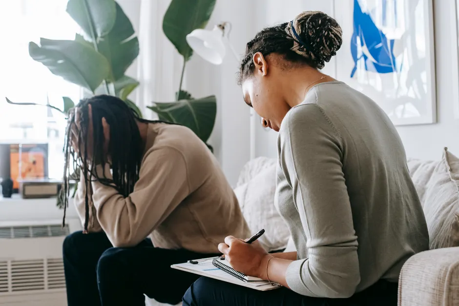 Professional female counselor taking notes in notepad while listening to depressed African American male client touching head in dismay in modern psychology center