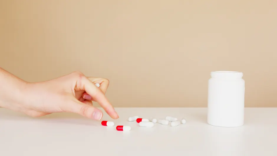 Side view of crop anonymous patient picking up one pill from pile of double colored pills scattered on table near white bottle