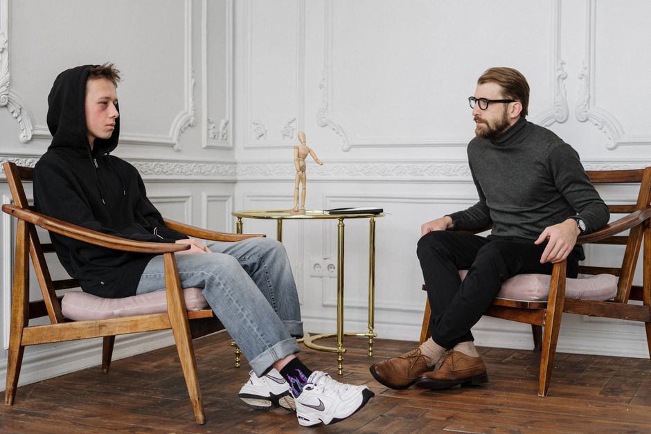 Teen boy in a hoodie having a conversation with a therapist in a stylish, minimalist room setting.