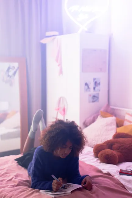 Teen girl with afro hair writing on bed in a cozy, pastel-colored bedroom with soft lighting.