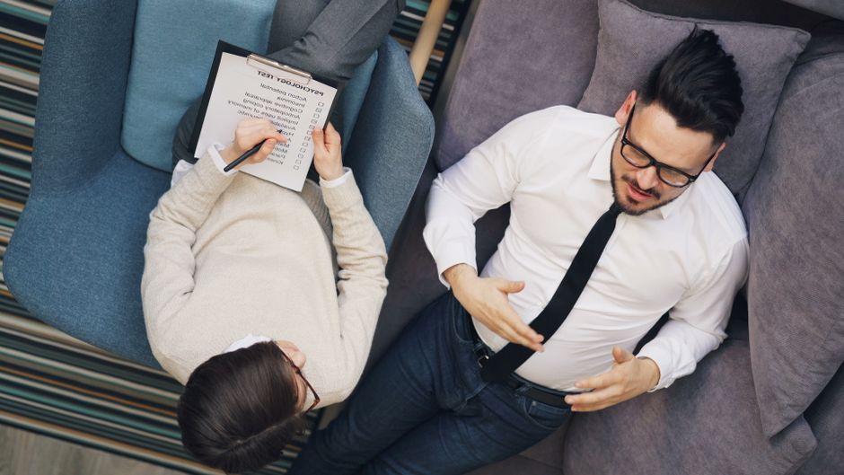 Therapist takes notes while patient discusses problems on a couch during a session.