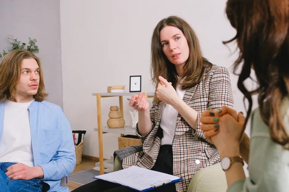 Thoughtful woman psychologist in stylish clothes siting with clipboard and talking with couple in light room
