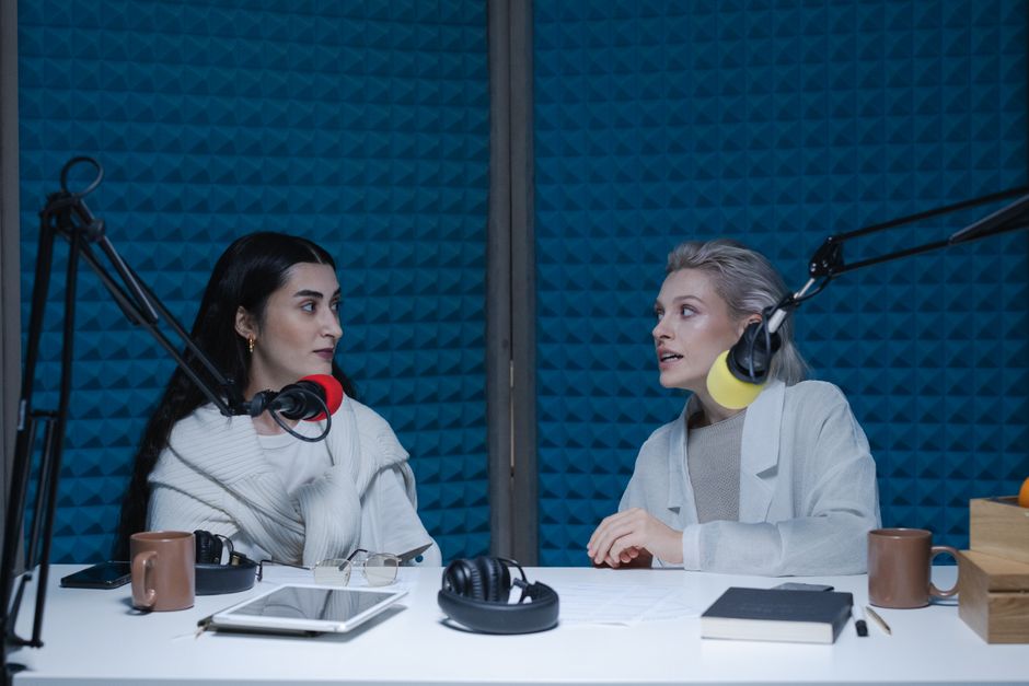 Two women engaged in conversation, recording podcast in studio with microphones and headphones.