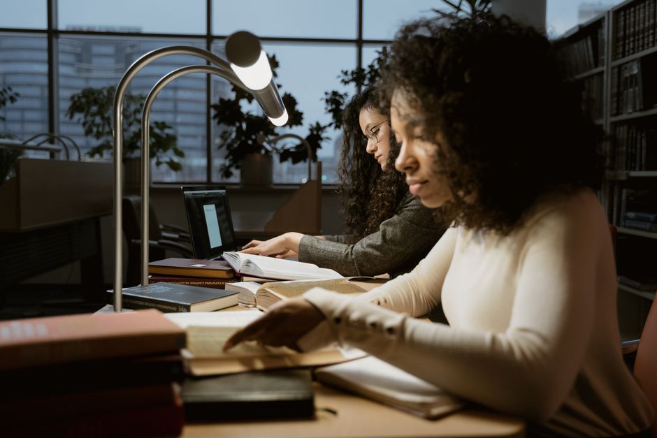 Two women intensely studying in a library setting, under warm desk lamps.