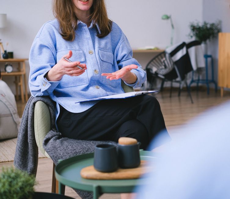 Unrecognizable professional female psychotherapist in chair with clipboard explaining solution of problem to faceless patient during psychotherapy session in office