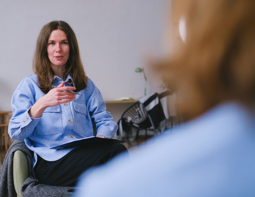 Woman in a blue jacket sitting indoors, engaged in conversation with another person.
