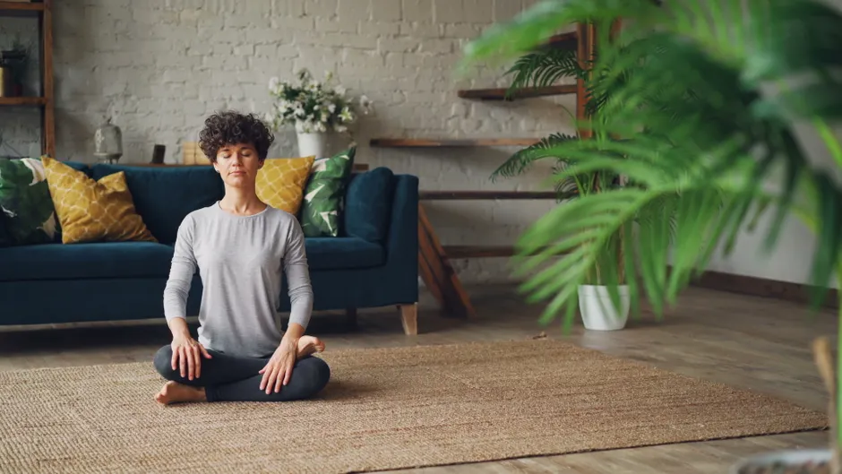 Woman meditating in a cozy living room with natural decor, promoting relaxation.