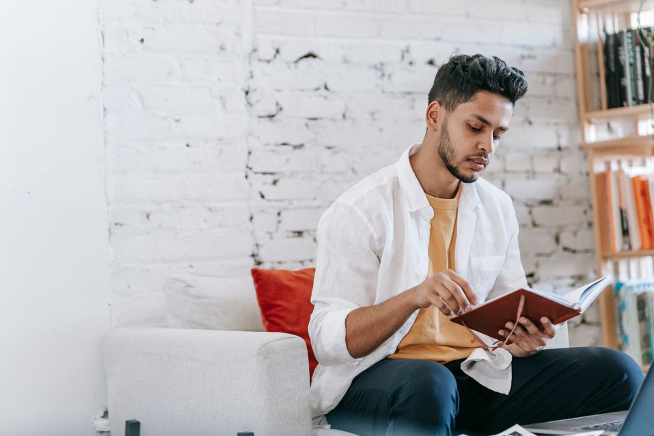 Young focused ethnic male wearing casual clothes reading notes in diary and sitting on comfortable armchair in light living room