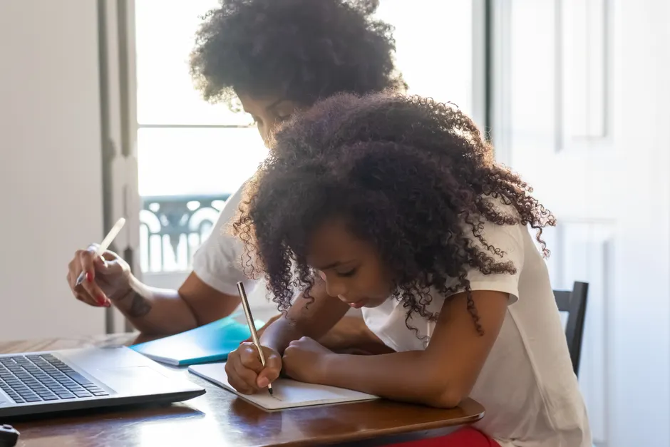 Young girl with afro hair studying with parent at home, focused on writing tasks.