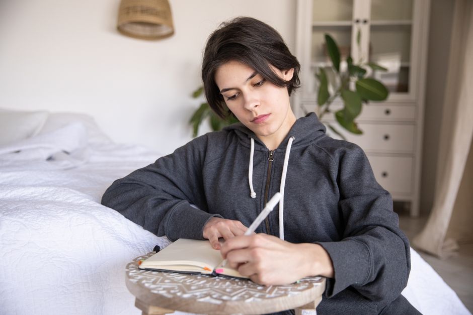 Young woman in a hoodie writing in a notebook at home, capturing a reflective moment.