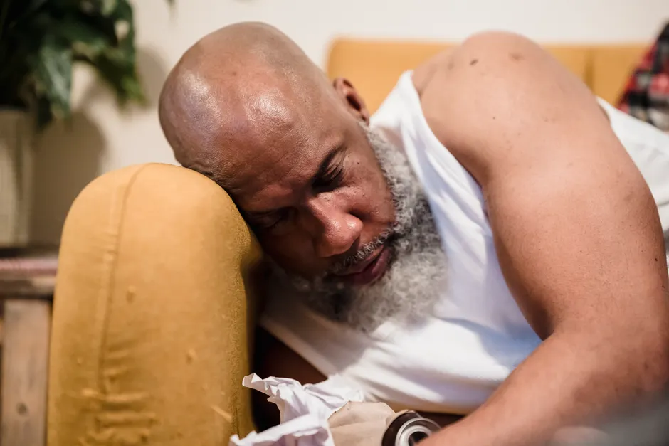 A bearded man sleeps on a couch, appearing tired in a cozy indoor setting.