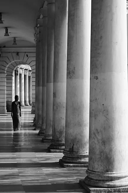 A black-and-white photo of a person walking through a column-lined hallway.