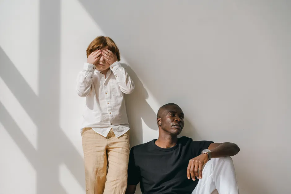 A child playfully covers their face while an adult sits calmly, bathed in warm sunlight.