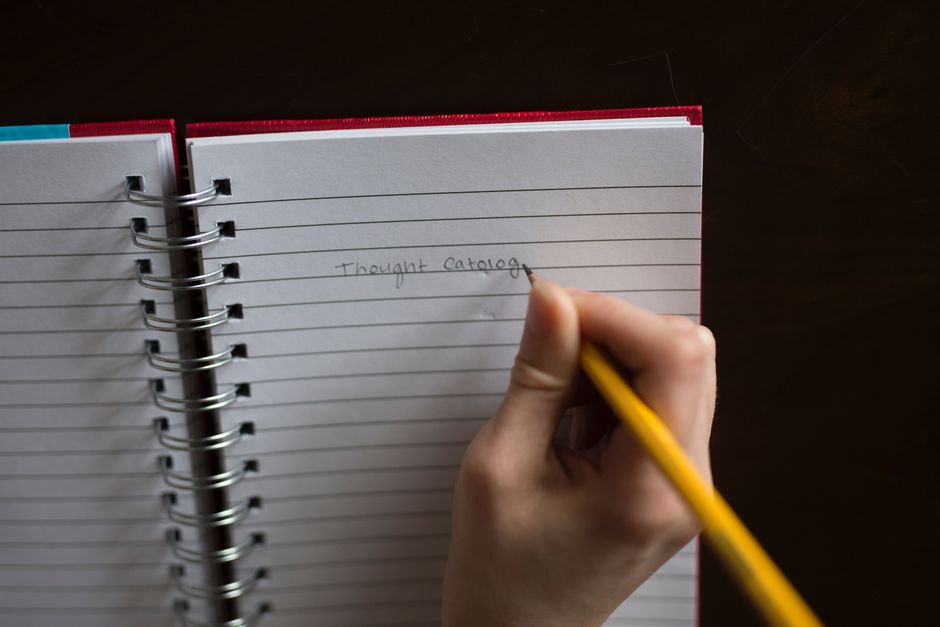 A close-up of a hand using a pencil to write in a spiral notebook with lined pages.