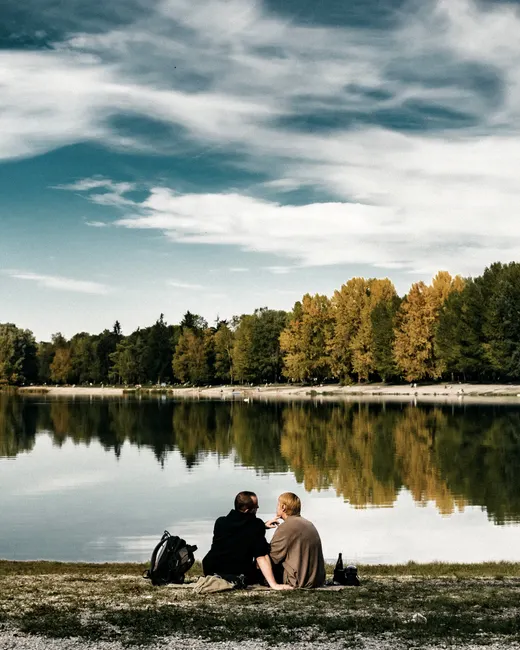 A couple sits by a serene lake, reflecting trees under a blue sky, enjoying nature.