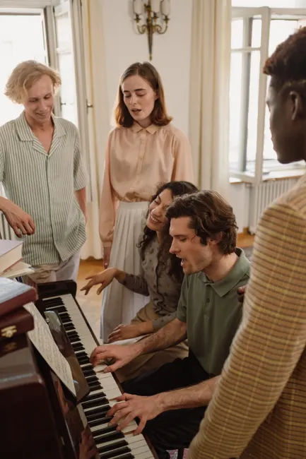 A diverse group of friends gathers around a piano, playing and singing together indoors.