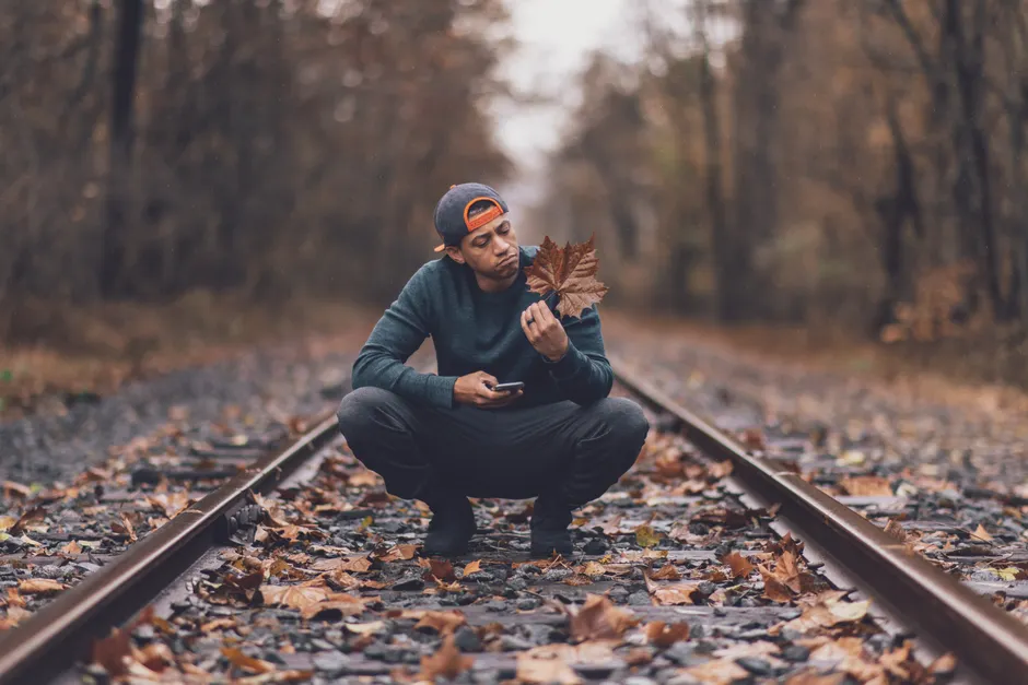 A man crouches on railroad tracks, holding a leaf, enjoying the autumn scenery.