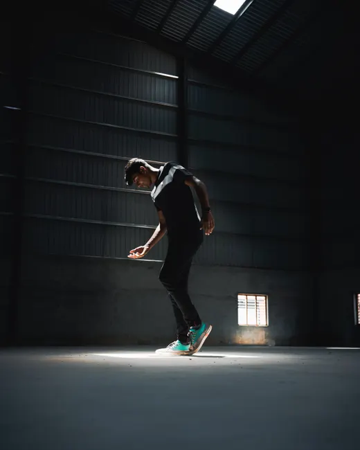 A man dances in a dramatic spotlight inside an industrial warehouse, showcasing youthful energy.