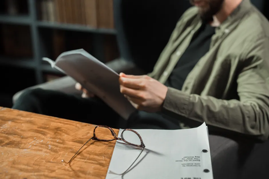 A man reading in a library with glasses and papers on a wooden table.