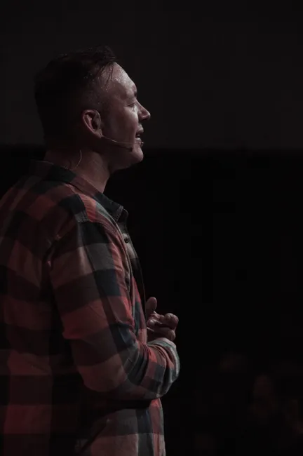 A man wearing a plaid shirt speaks into a headset microphone on a dimly lit stage.