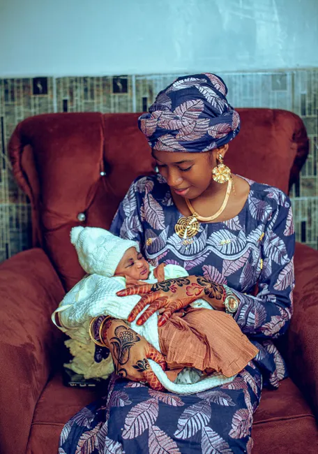 A mother lovingly holds her newborn baby, both in traditional African attire, seated indoors.