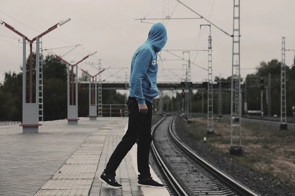 A person in a blue hoodie stands on a railway platform, contemplating near the tracks.