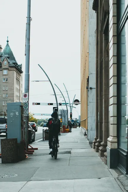 A person rides a bicycle along an urban sidewalk, surrounded by tall buildings and streetlights.