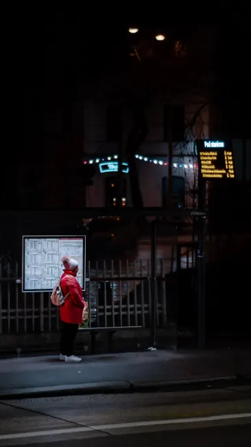 A person waits alone at a bus stop in the city at night, under streetlights.