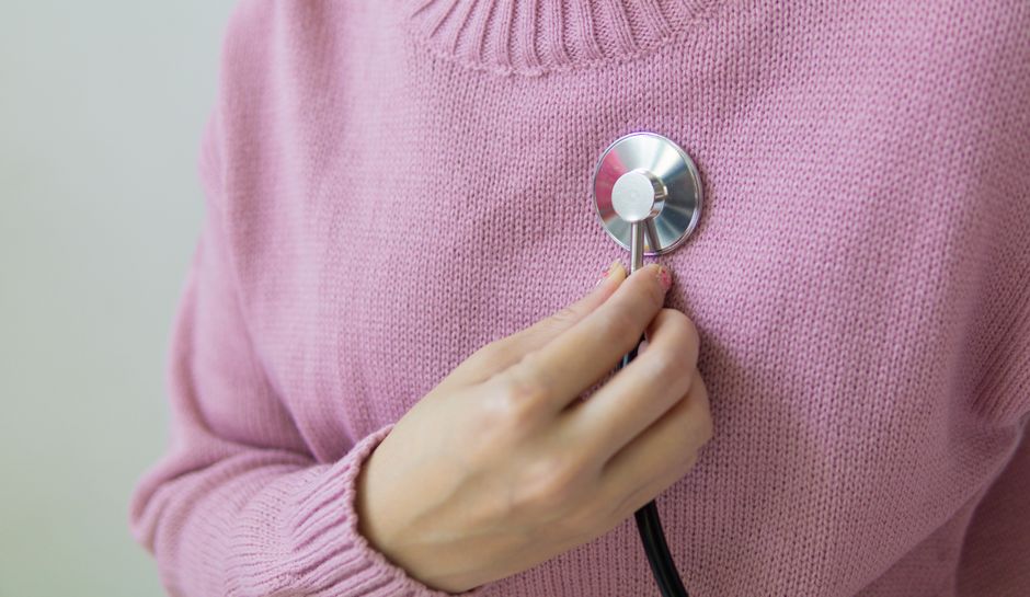 A person wearing a pink sweater holds a stethoscope to their chest for a health check.