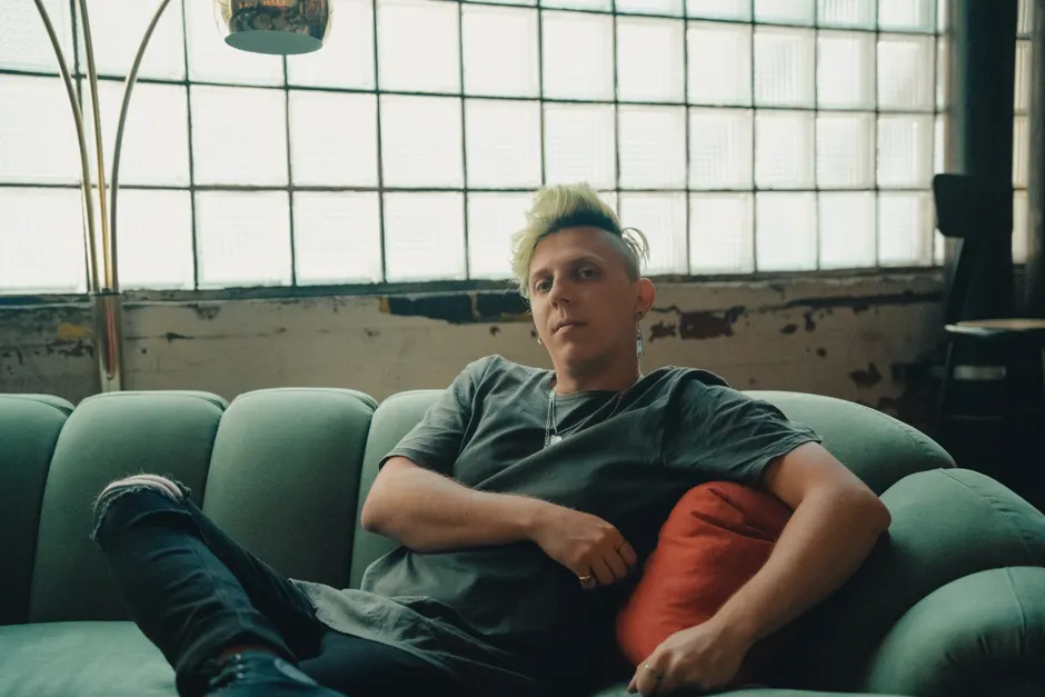 A person with green hair relaxing on a teal sofa in a chic industrial indoor setting.