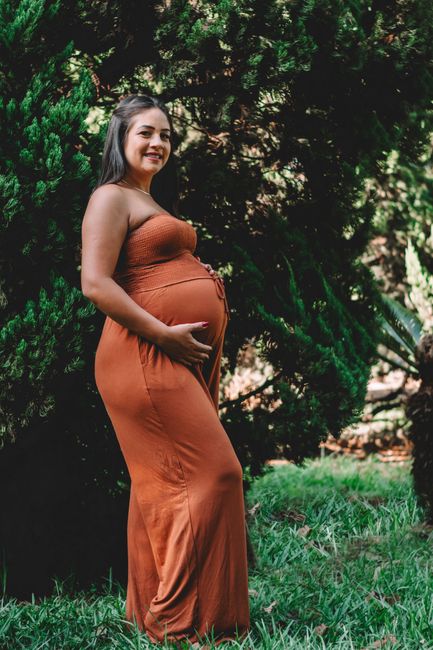 A pregnant woman standing and smiling in a lush outdoor setting, captured in a warm portrait.