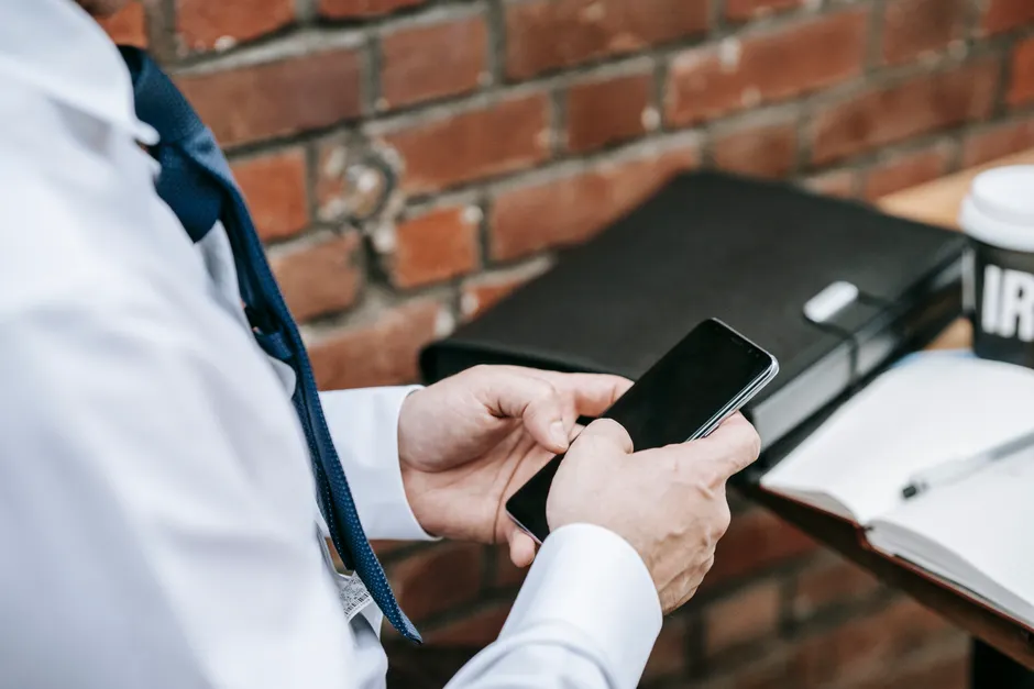 A professional in corporate attire using a smartphone against a brick wall indoors.