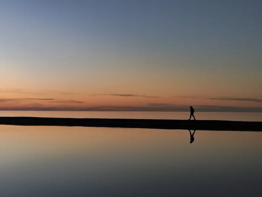A serene sunset reflection scene with a person walking along the beach, creating a silhouette.
