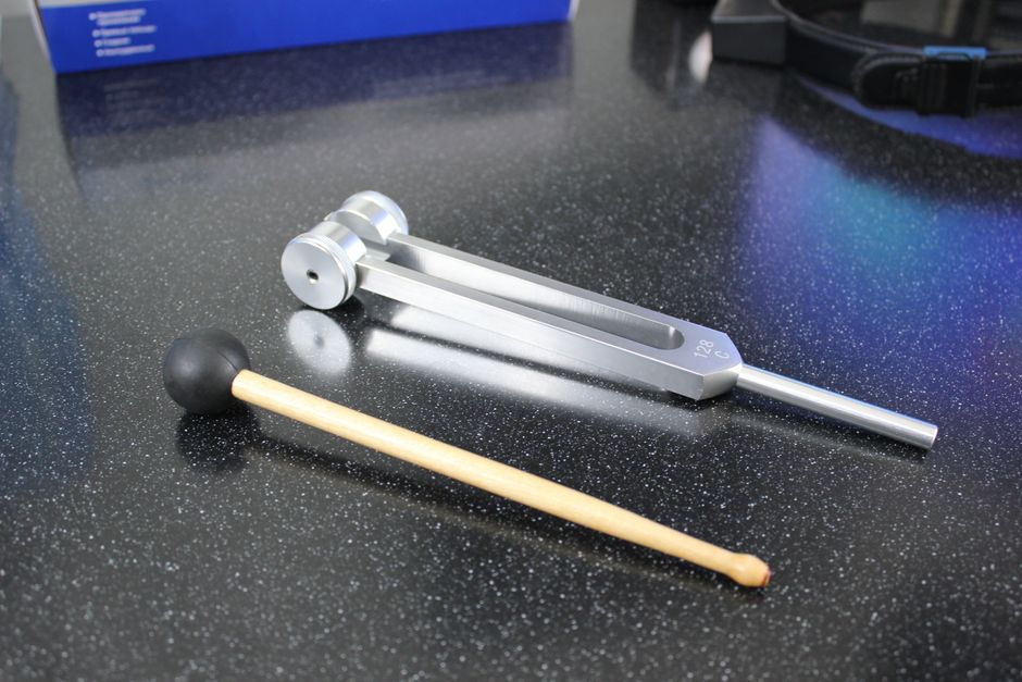 A shiny metal tuning fork with mallet on speckled black table, for sound therapy or music.