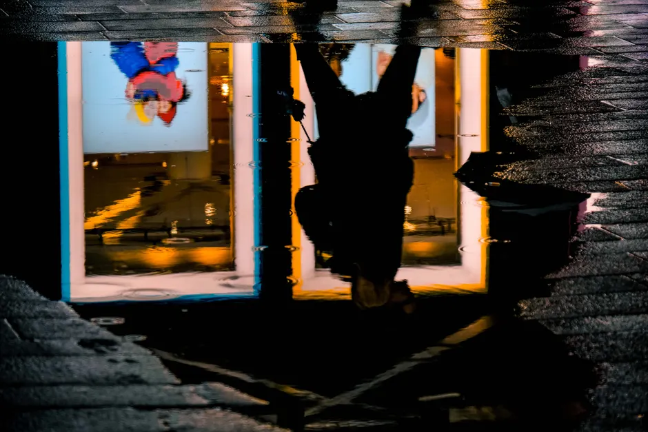 A silhouette of a person is reflected in a water puddle, capturing urban lights and textures.