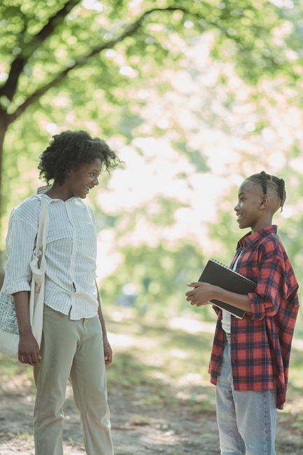 A smiling teenager talks with an adult outdoors, holding a notebook in a sunny park.