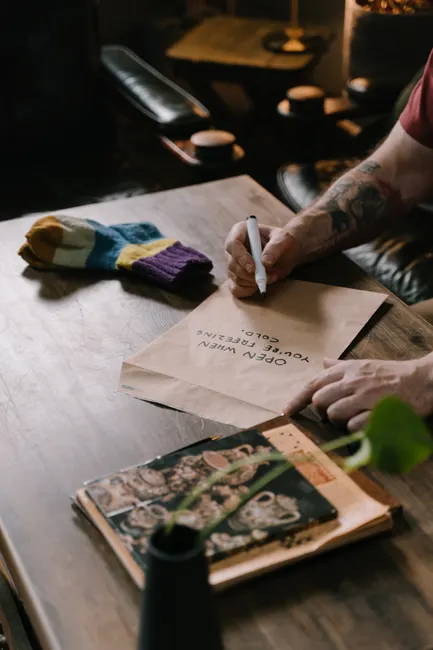 A tattooed person writes a message on an envelope beside colorful socks on a wooden table.