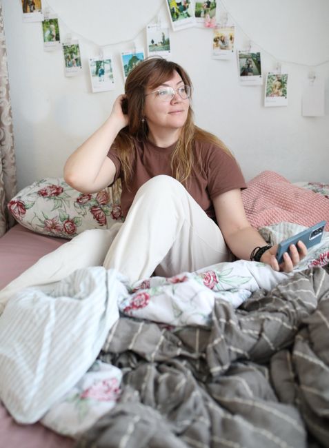 A woman comfortably sitting in bed holding a smartphone, surrounded by cozy decor.