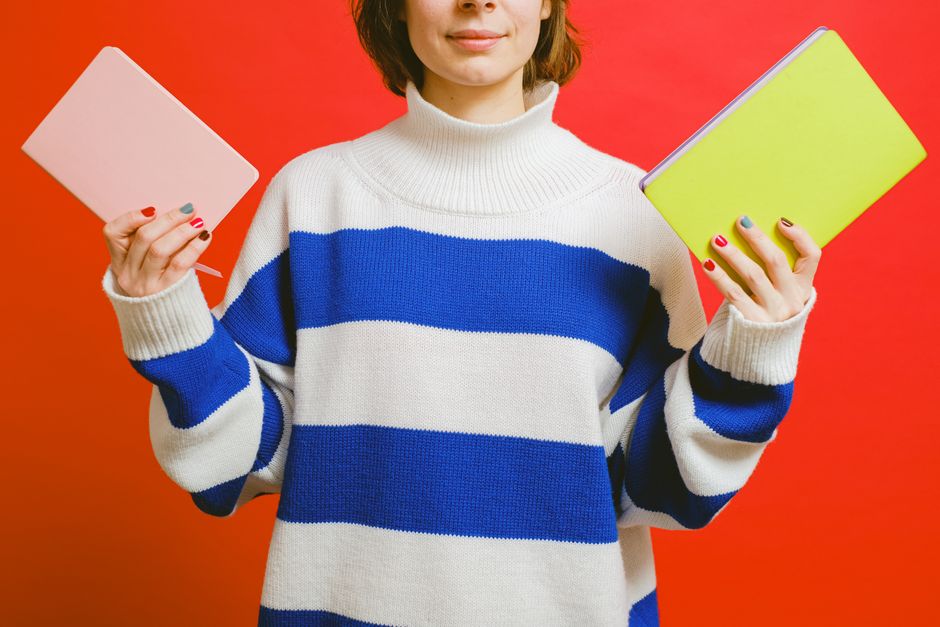 A woman holds pink and yellow notebooks while wearing a white and blue striped sweater against a red background.