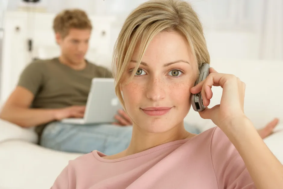 A woman smiling while talking on her mobile phone, with a man focused on his laptop in the background.