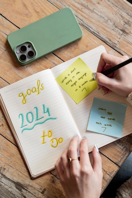 A woman writes goals in a notebook with a smartphone and sticky notes nearby.