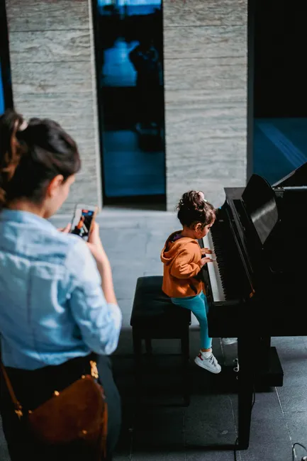 A young child plays the piano indoors while a parent records the moment using a cellphone.