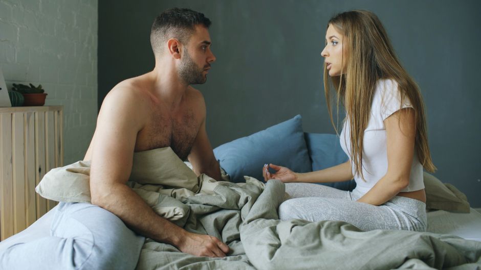 A young couple discusses something important while sitting on their bed in a cozy room.