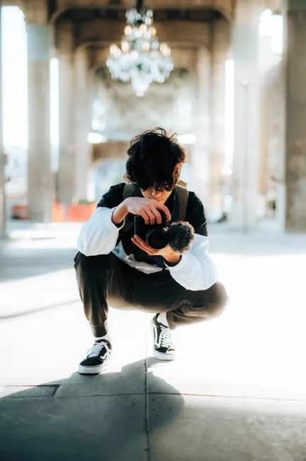 A young man squats under a chandelier focusing on his camera in an urban setting.