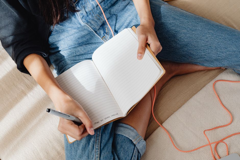 A young woman sitting comfortably, writing in a notebook while listening to music.