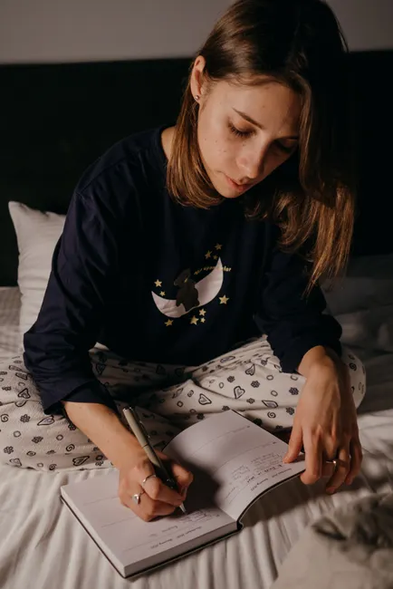 A young woman writing in a journal while sitting on her bed, captured in an intimate and relaxed atmosphere.