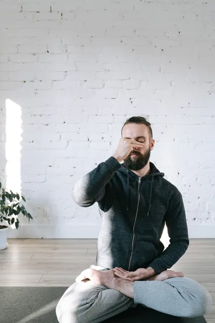 Adult man in yoga pose practicing breathing exercise indoors against white brick wall.